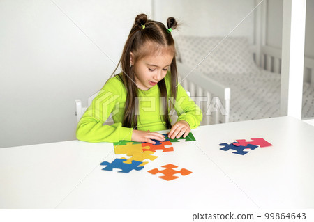 A girl in a bright light green T-shirt is sitting at a desk in the children's room, putting the colored puzzle pieces together. 99864643