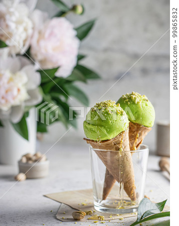 Two pistachio ice cream cones in a glass with chopped pistachios and flowers in the back Two pistachio ice cream cones in a glass with chopped pistachios and flowers in the back 99865832