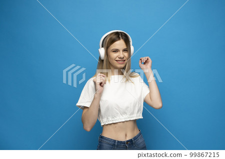 Cheerful young pretty girl smiling while listening music in headphones and dancing on blue background. 99867215