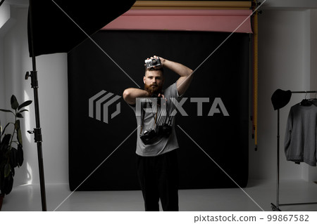 Bearded professional photographer in a grey t-shirt with a bunch of different cameras in a hands and on a shoulder looking on a camera and ready for make a good shoot. Bearded professional photographer in a grey t-shirt with a bunch of different cameras in a hands and on a shoulder looking on a camera and ready for make a good shoot. 99867582