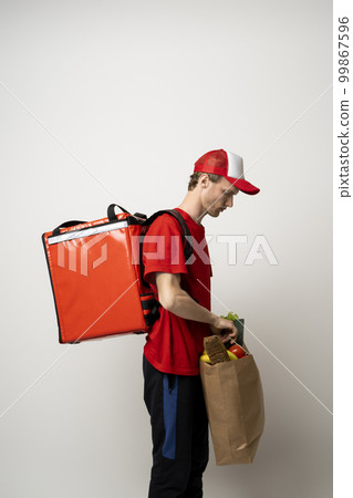 Young man in a red t-shirt and a cap holding paper bag with products on white background. Food delivery service. 99867596
