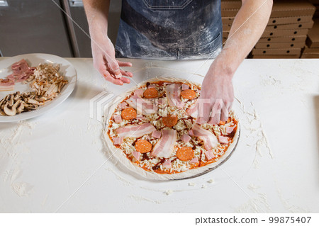 Cooking pizza. Lays out the cheese and salami filling, ingredients on the dough preform. Closeup hand of chef baker in uniform white apron cook at kitchen 99875407