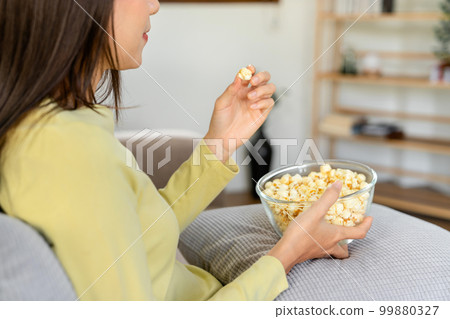 Young asian woman putting a bowl of popcorn on pillow and eating popcorn while sitting on the big comfortable Young asian woman putting a bowl of popcorn on pillow and eating popcorn while sitting on the big comfortable 99880327