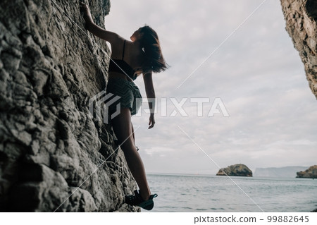 Sports Woman Climbing The Rock. Young woman With slim fit body climbing in volcanic basalt cave with beautiful sea view. The athlete girl trains in nature. Woman overcomes difficult climbing route. 99882645