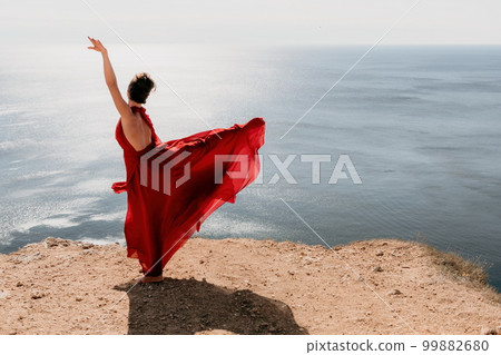 Woman in red dress on sea. Side view a Young beautiful sensual woman in a red long dress posing on a rock high above the sea on sunset. Girl on the nature on blue sky background. Fashion photo. 99882680