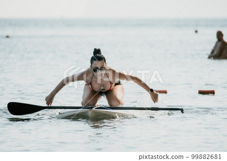 Sea woman sup. Silhouette of happy middle aged tanned woman in rainbow bikini, surfing on SUP board, confident paddling through water surface. Idyllic sunset. Active lifestyle at sea or river. 99882681