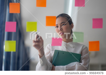 A businesswoman uses Post-it notes to share ideas, showcasing the concept of brainstorming. The sticky notes are placed on a glass wall. 99884978