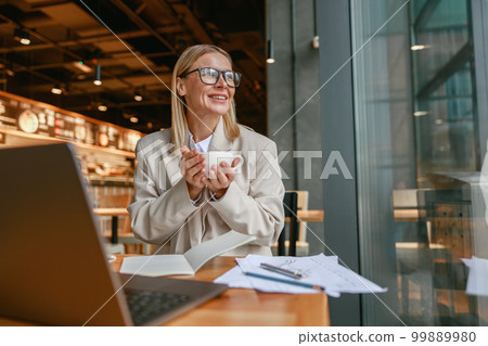 Attractive businesswoman holding cup of coffee while sitting in cafe during working day Attractive businesswoman holding cup of coffee while sitting in cafe during working day 99889980