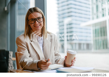 Smiling businesswoman in glasses drinking coffee and making notes while working in cafe 99890126