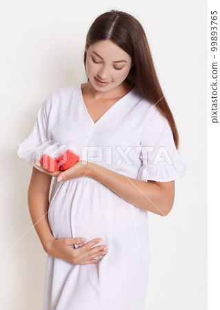 Gently pregnant woman with red booties, touching her belly, expectant mother with baby's booties posing against white wall, brunette lady with long beautiful expecting child. Gently pregnant woman with red booties, touching her belly, expectant mother with baby's booties posing against white wall, brunette lady with long beautiful expecting child. 99893765