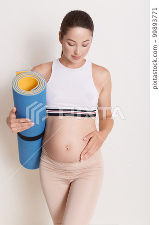Young attractive pregnant female touching her belly while holding yoga mat in hands, expectant mother doing physical exercises, posing isolated over white background. 99893771