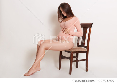 Attractive beautiful pregnant female wearing powder color combidress sitting on brown wooden chair and looking on her belly, woman with long brown hair. Attractive beautiful pregnant female wearing powder color combidress sitting on brown wooden chair and looking on her belly, woman with long brown hair. 99893809