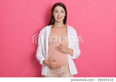 Young attractive pregnant fashion woman wearing casual attire, looking directly at camera, having positive emotions. camera, touching her belly with both hands against rose background. Young attractive pregnant fashion woman wearing casual attire, looking directly at camera, having positive emotions. camera, touching her belly with both hands against rose background. 99893818