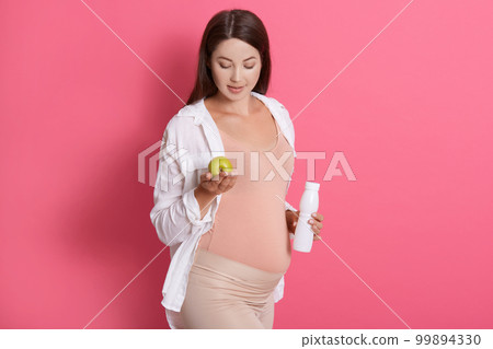 Pregnant young woman holding green apple and bottle of yogurt or milk. Healthy food during pregnancy. Expectant mother against pink wall, wearing casual clothing. Pregnant young woman holding green apple and bottle of yogurt or milk. Healthy food during pregnancy. Expectant mother against pink wall, wearing casual clothing. 99894330