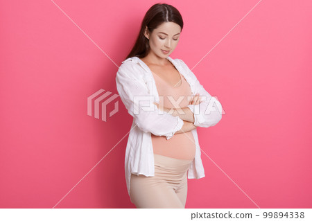 Serious and confident pregnant woman with arms crossed looking down on her belly, wearing stylish clothing, dark haired expectant mother posing isolated over rose background. Serious and confident pregnant woman with arms crossed looking down on her belly, wearing stylish clothing, dark haired expectant mother posing isolated over rose background. 99894338