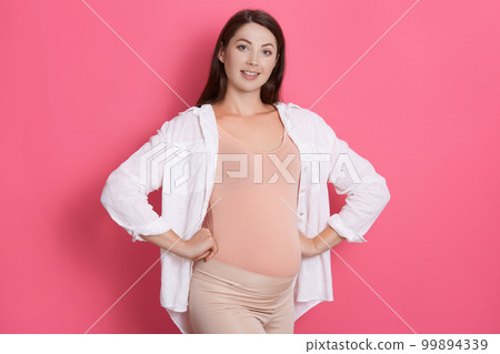 Happy healthy young pregnant woman standing with hands on hips and looking at camera with beaming smile, dark haired future mommy posing isolated over rosy background. Happy healthy young pregnant woman standing with hands on hips and looking at camera with beaming smile, dark haired future mommy posing isolated over rosy background. 99894339