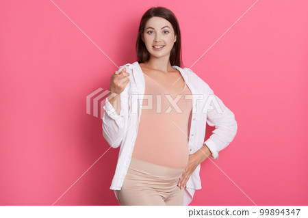 Happy smiling pregnant lady pointing at camera, looks happy, touches her belly, wearing stylish clothing against pink wall, dark haired expectant mother. 99894347