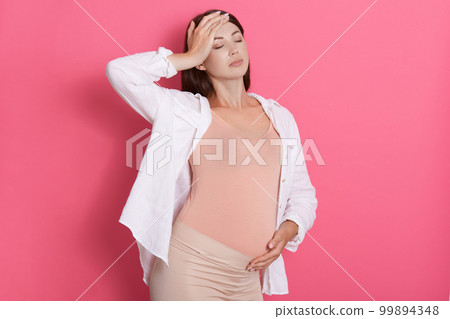 Young pregnant woman feeling sick, keeps eyes closed, touching her head and belly, suffering from headache and toxicosis, posing isolated over pink background. 99894348