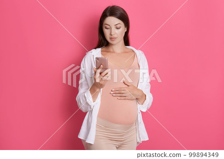 Pretty pregnant woman using her smart phone and touching her belly, looking at device's screen, looks concentrated, posing isolated over rosy background. 99894349