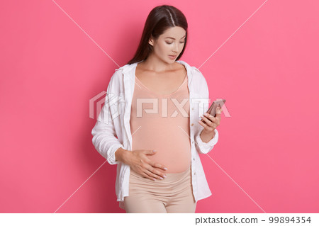 Young pregnant woman with dark hair wearing white shirt holds smart phone in her hand and looks at camera isolated over pink background, texting with doctor and touches belly. 99894354