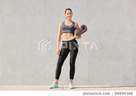 Full length portarait of beautiful woman with healthy skin, pony tail, has workout, dressed in sportsclothes, poses against grey background. Athletic sportswoman ready for yoga class. Copy space. 99894366
