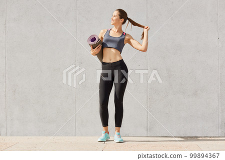 Full length photo of healthy young woman in sportswear holding yoga mat, touches her ponytail, be photographed after training, model posing solated over gray background. Copy space for advertisment. Full length photo of healthy young woman in sportswear holding yoga mat, touches her ponytail, be photographed after training, model posing solated over gray background. Copy space for advertisment. 99894367