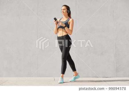 Thoughtful pleasant looking sportswoman in top, leggings and sportshoes, listens favourite songs in headphones, holds modern mobile phone, has training early in mornning. Sport and technology 99894378