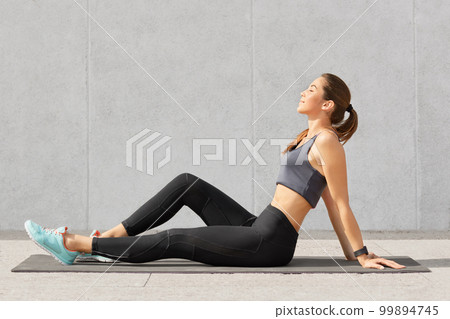 People, sport and relaxation concept. Relaxed fitness woman with perfect figure sits on exercise mat, keeps eyes closed, dreams about something after having morning workout or yoga practice. 99894745