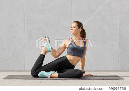 People and active exercises concept. Beautiful European young woman with darkhair, dressed in sportswear, raises legs for stretching before aerobics, poses on fitness mat, isolated on grey wall 99894766