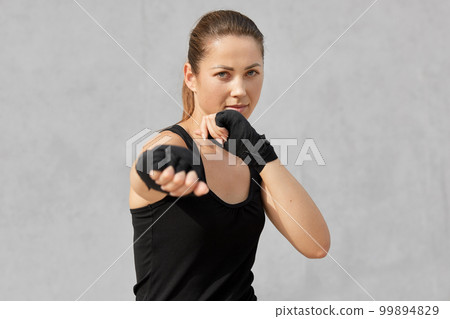 Photo of female boxer in defensive stance, looks fierce, dressed in black t shirt, bandages on hands, ready to fight with opponent, stands against over grey background. People and boxing concept Photo of female boxer in defensive stance, looks fierce, dressed in black t shirt, bandages on hands, ready to fight with opponent, stands against over grey background. People and boxing concept 99894829