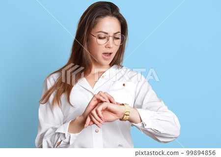 Closeup portrait of beautiful business woman being late for meeting . Young dark haired stressed attractive girl checks time with opened mouth, wears white t shirt isolated over blue background. Closeup portrait of beautiful business woman being late for meeting . Young dark haired stressed attractive girl checks time with opened mouth, wears white t shirt isolated over blue background. 99894856