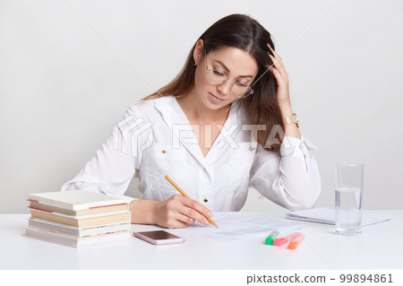 Busy dark haired woman makes notes, underlines information in papers, wears white blouse, spectacles, surrounded with pile of books, smart phone, glass of water, isolated over white studo wall 99894861