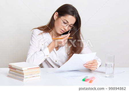 Photo of young businesswoman works remotely, talks on mobile phone, checks bank balance, holds pencil, verifies documents, uses scientific liteature for writing course paper, wears formal clothes 99894862