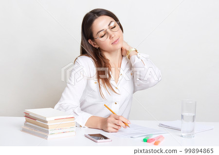 Image of busy woman keeps hand on neck, feels tired after long work with documents, sits at workplace, writes creative ideas for creating publication, has shut eyes, waits for call on cell phone 99894865