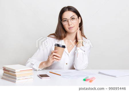 Indoor shot of pleasant looking Caucasian woman holds takeaway coffee, dressed in white shirt, looks directly at camera, has break after paper work. Female teacher prepares for classes indoor 99895446