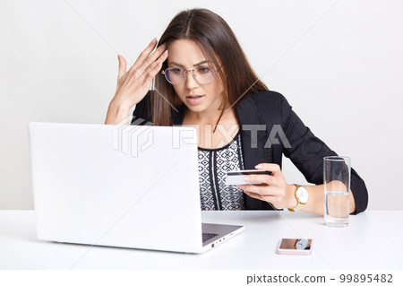 Surprised woman with dissatisfied expression, makes online transaction, works with laptop computer, makes shopping in web store, uses credit card, drinks glass of water, isolated on white wall 99895482