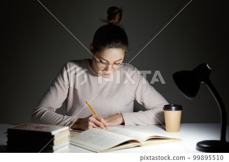 Photo of young female writes something with pensil, uses table lamp, prepares for examination, sits at desktop, wears casual shirt and optical glasses, poses against dark background. Study concept Photo of young female writes something with pensil, uses table lamp, prepares for examination, sits at desktop, wears casual shirt and optical glasses, poses against dark background. Study concept 99895510