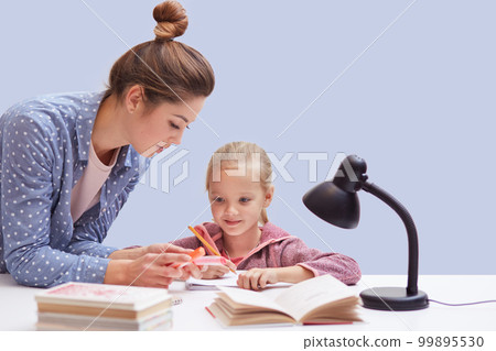 Studio shot of little charming girl sits at table, has difficult homework task, her mother trying to help daughter and explains mathematics rules, uses reading lamp for good vision. Education concept. 99895530