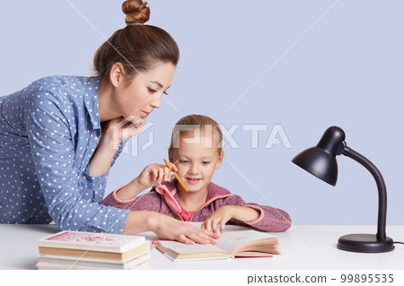 Close up portrait of beautiful Caucasian woman helping her doughter to do school homework, female with bunch on head, wearing casual outfit, little cute girl sitting at table surrounded by books. 99895535