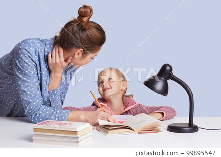 Image of mother and daughter sitting at table surrounded by books looking at each other with love, doing homework together, mummy helps little girl to do sums. Children, school, education concept. Image of mother and daughter sitting at table surrounded by books looking at each other with love, doing homework together, mummy helps little girl to do sums. Children, school, education concept. 99895542