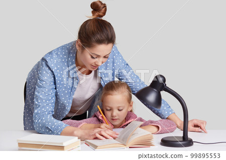 Horizontal shot of experienced young mother leans near her small child, helps to do home assignment, shows what to rewrite in book, surrounded with reading lamp, isolated over white background Horizontal shot of experienced young mother leans near her small child, helps to do home assignment, shows what to rewrite in book, surrounded with reading lamp, isolated over white background 99895553