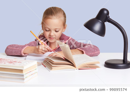 Close up portrait of little charming girl sits at white desk, does homework task, try to write composition or does sums, looks concentrated, uses reading lamp for good vision, isolated over blue wall. Close up portrait of little charming girl sits at white desk, does homework task, try to write composition or does sums, looks concentrated, uses reading lamp for good vision, isolated over blue wall. 99895574