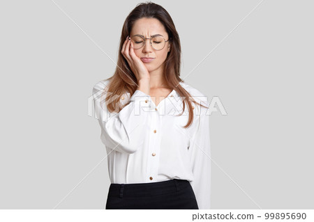 Photo of tired overworked female office worker keeps eyes closed, feels sleepy, holds hand on cheek, dressed in stylish clothes, isolated over white background. Fatigue businesswoman has toothache 99895690