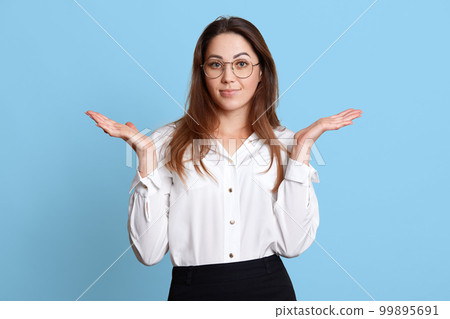 Helpless charismatic young female with long hair making gesture, raising her hands, looks confused about work, posing isolated over light blue background in studio. People and office work concept. Helpless charismatic young female with long hair making gesture, raising her hands, looks confused about work, posing isolated over light blue background in studio. People and office work concept. 99895691