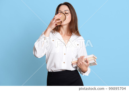 Slender smart young female drinks coffee with pleasure during break, holds bunch of textbooks, being in hurry. Good looking business wears formal white blouse and black skirt. Copy space for ad. Slender smart young female drinks coffee with pleasure during break, holds bunch of textbooks, being in hurry. Good looking business wears formal white blouse and black skirt. Copy space for ad. 99895704