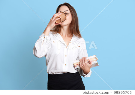 Indoor studio shot of slender hard working office worker having coffee break, drinking hot drink from papercup, holding bunch of books in one hand, posing isolated over light blue background. 99895705