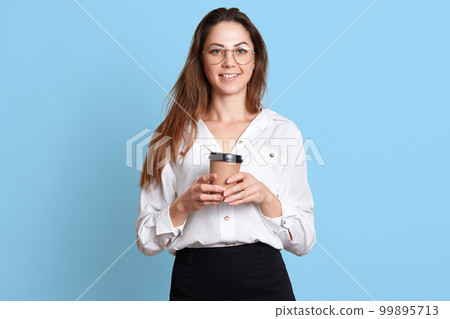 Smiling energetic young girl enjoys drinking hot coffee, holds paper cup in hands, poses, wears formal black skirt and white blouse, takes rest after hard working day. Copy space for advertisement. Smiling energetic young girl enjoys drinking hot coffee, holds paper cup in hands, poses, wears formal black skirt and white blouse, takes rest after hard working day. Copy space for advertisement. 99895713