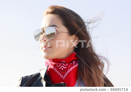 Close up portrait of beautiful girl wearing leather jacket, red bandana and cool sunglasses. Female person posing isolated over sky, wind blowing on her long dark hair, lady looks thoughtfully aside. 99895719