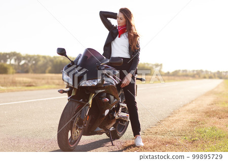 Horizontal shot of active female motorcyclist pose on fast bike in open air, wears red bandana, black leather jacket and sneakers, focused aside, takes break on empty road. People and leisure concept Horizontal shot of active female motorcyclist pose on fast bike in open air, wears red bandana, black leather jacket and sneakers, focused aside, takes break on empty road. People and leisure concept 99895729