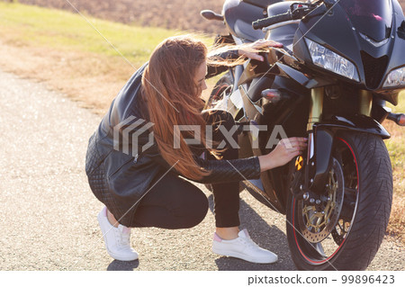 Image of female biker wears black clothes and white sneakers, repairs motorbike, stands on road, solves problem on road, likes journey. Outdoor shot of active motorcyclist repairs transport. 99896423